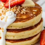 Stack of fluffy coconut cream pancakes topped with whipped cream and coconut flakes. Delicious and easy breakfast recipe.